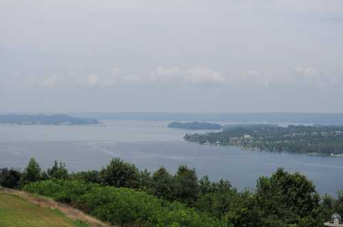 A scenic view of a calm lake surrounded by green trees and distant hills under a cloudy sky.