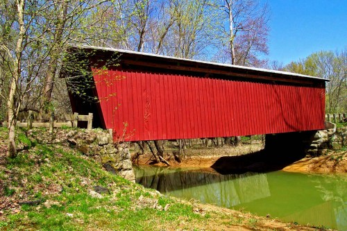 A red covered bridge spans a calm green river, surrounded by trees and a clear blue sky.