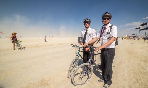Two men in suits and helmets stand with bicycles in a dusty landscape, holding a book, at a festival.