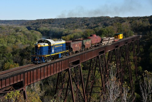 A blue and yellow train crosses a tall bridge over a lush green valley, with smoke billowing from its engine.