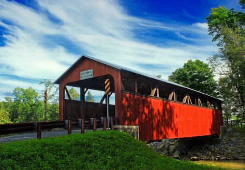 A red covered bridge spans a small stream, surrounded by green trees and a blue sky with wispy clouds.