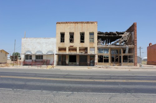 Abandoned buildings with crumbling facades and broken windows along a deserted road under a clear blue sky.
