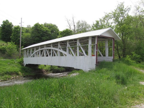 A white covered bridge spans a small stream, surrounded by lush green grass and trees under a clear sky.