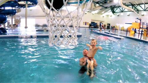 A child joyfully rides on an adult's shoulders in a swimming pool, with water slides and other kids in the background.