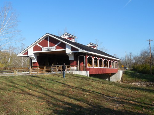A red and white covered bridge with a wooden walkway, surrounded by trees and a clear blue sky.