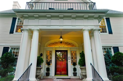 A grand entrance of a house featuring a red door, white columns, and decorative plants on either side.