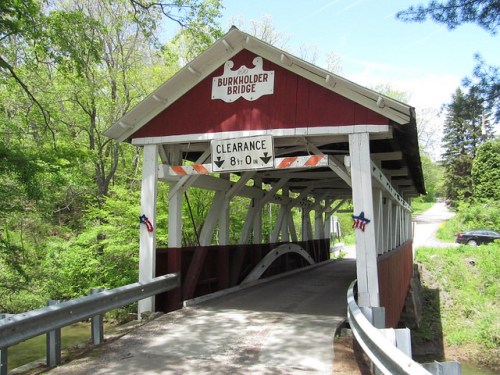 A red covered bridge with a sign indicating clearance, surrounded by lush green trees and a winding road.