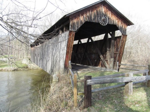 A rustic covered bridge made of wood spans a calm river, surrounded by trees and grassy areas.