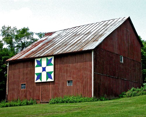 A rustic red barn with a colorful quilt pattern on its side, surrounded by green grass and trees.