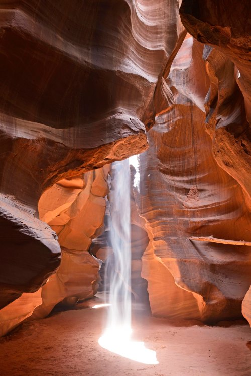 A narrow canyon with smooth, wavy walls and a beam of light shining down onto the sandy floor.