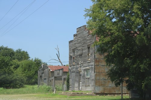 Abandoned buildings with weathered wood and rusted metal, surrounded by overgrown grass and trees under a clear blue sky.
