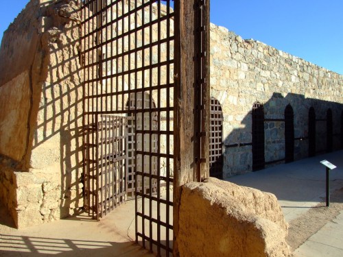 A weathered stone structure with a barred gate, showcasing remnants of an old building under a clear blue sky.