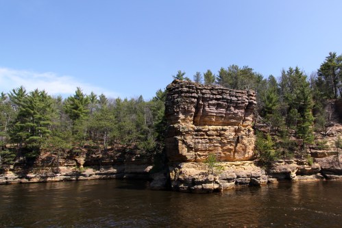 A rocky cliff rises above a calm river, surrounded by lush green trees under a clear blue sky.