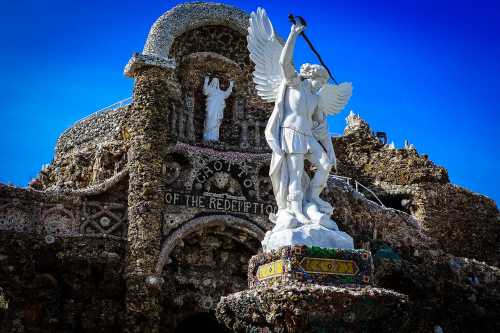 A white angel statue stands in front of a stone structure with intricate designs and a blue sky above.