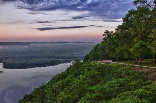 A serene landscape at dawn, featuring a river, lush greenery, and a misty horizon under a colorful sky.