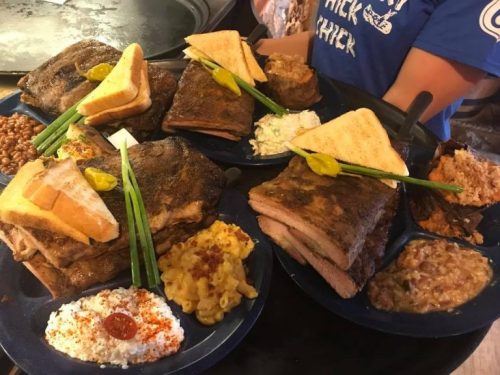 A variety of barbecue dishes on blue plates, including meats, sides, and garnishes like pickles and green onions.