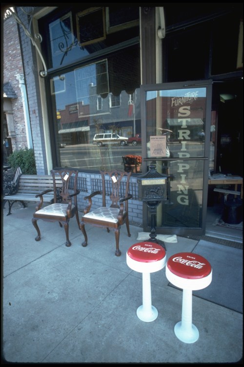 Two vintage chairs and Coca-Cola stools outside a furniture store, with a view of the street in the background.