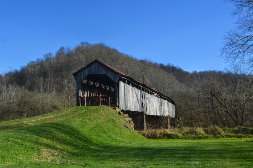 A rustic covered bridge stands on a grassy hill, surrounded by trees and rolling hills under a clear blue sky.