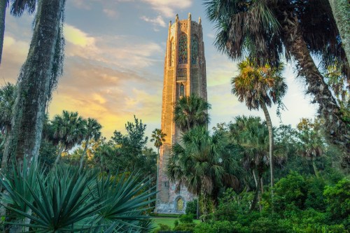 A tall stone tower surrounded by lush greenery and palm trees under a colorful sky at sunset.