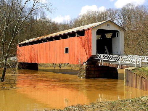 A red covered bridge partially submerged in brown floodwaters, surrounded by trees under a blue sky.