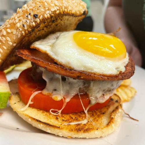 A close-up of a sesame seed bun sandwich with a fried egg, bacon, tomato, and melted cheese.