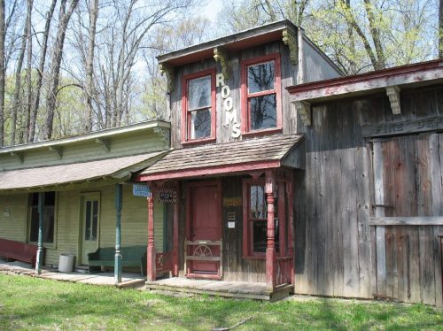 A rustic, two-story building with a sign reading "ROOMS," surrounded by trees and older wooden structures.