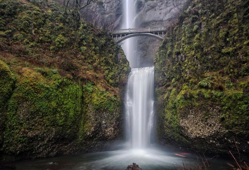 A majestic waterfall cascades down rocky cliffs, with a stone bridge arching above, surrounded by lush greenery.