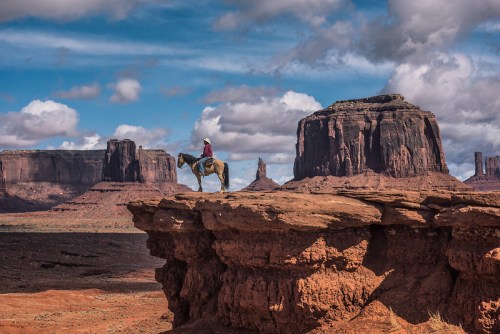 A rider on horseback stands on a rocky outcrop, with dramatic cliffs and clouds in the background.