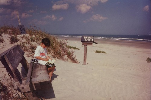A boy sits on a bench by the beach, writing near a sign that reads "Kindred Spirit," with ocean waves in the background.