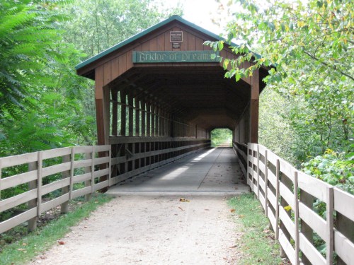 A wooden covered bridge with a sign reading "Bridge of Dreams," surrounded by trees and a gravel path.