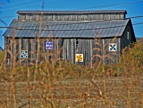 A weathered barn with colorful quilt patterns on its walls, surrounded by tall grass and a clear blue sky.