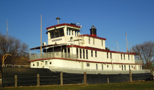 A large white and red tugboat docked on land, with a clear blue sky and trees in the background.