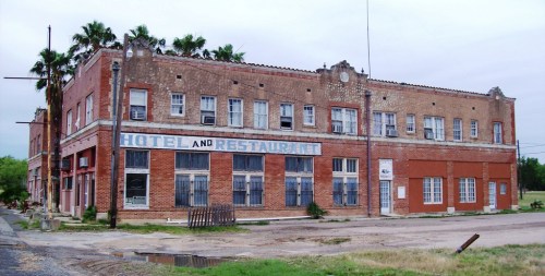 Abandoned hotel and restaurant building with a brick facade, palm trees, and boarded windows. Overcast sky.
