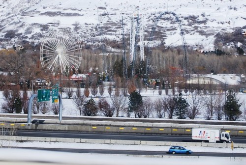 A snowy landscape featuring a ferris wheel, roller coaster, and highway with vehicles in the foreground.