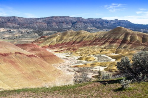 Colorful, layered hills of the Painted Hills in Oregon, with a clear blue sky and distant mountains in the background.