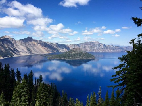 A serene view of Crater Lake, featuring a blue lake, an island, and surrounding mountains under a partly cloudy sky.