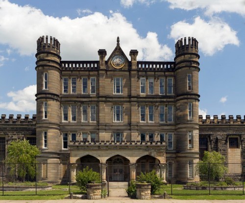 Historic stone building with towers and a clock, set against a blue sky with clouds. Lush greenery in the foreground.
