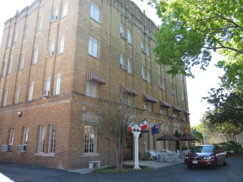 A brick building with awnings, featuring flags and outdoor seating, surrounded by trees and a parked car.