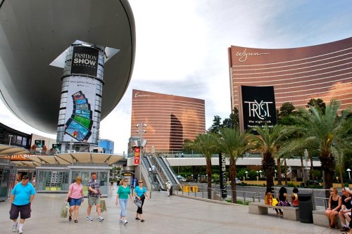 People walking in a shopping area with large billboards and modern buildings in Las Vegas. Palm trees in the foreground.