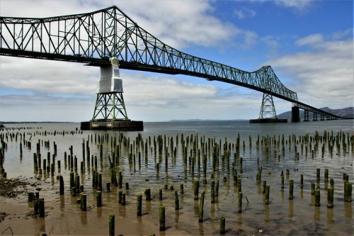 A large metal bridge spans over water, with wooden pilings visible in the foreground and a cloudy blue sky above.