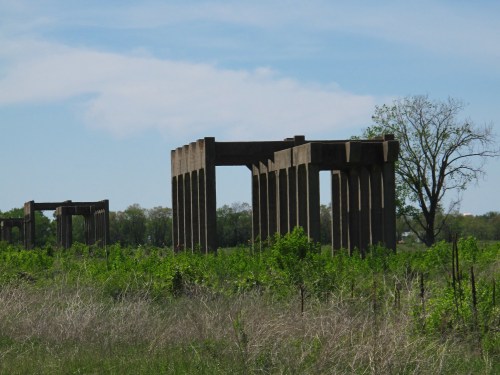 Abandoned concrete structures rise amidst tall grass and greenery under a clear blue sky.