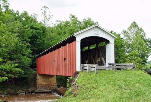 A red and white covered bridge spans a small stream, surrounded by lush green trees and grass.