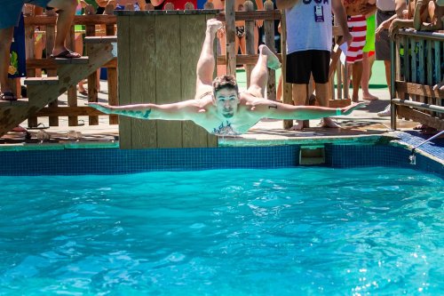 A person diving into a pool from a wooden platform, surrounded by onlookers enjoying a sunny day.