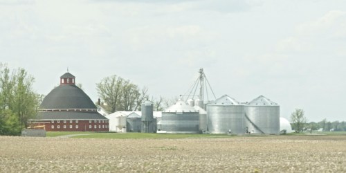A rural farm scene featuring a round red barn and several silos against a cloudy sky. Fields in the foreground.