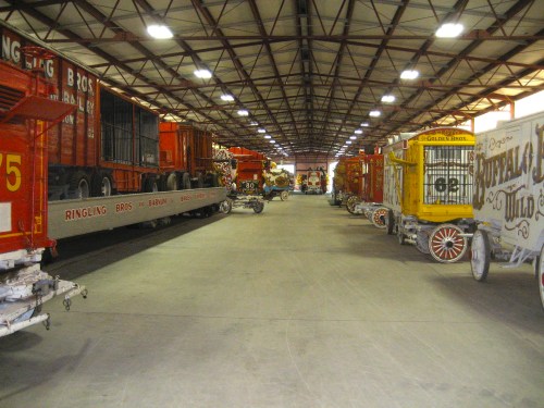 Interior of a large warehouse displaying colorful vintage circus wagons and trucks, lined up in rows.