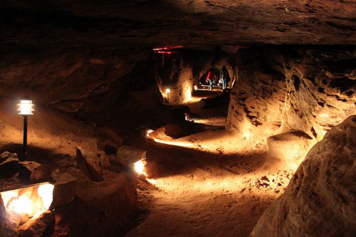 Dimly lit cave interior with sandy paths and glowing lights, showcasing rock formations and visitors in the background.