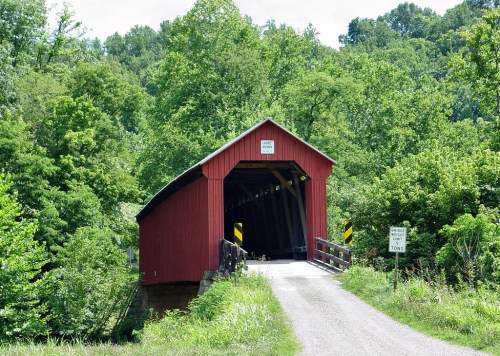 A red covered bridge surrounded by lush green trees, leading to a gravel road with a sign nearby.