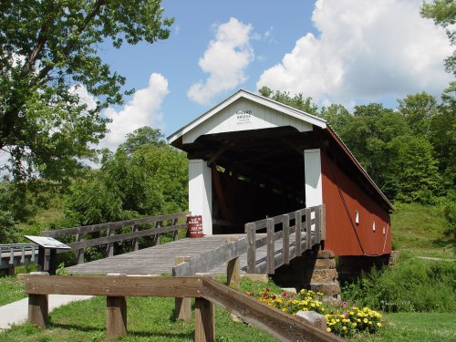 A red covered bridge spans a small stream, surrounded by green trees and blue skies with fluffy clouds.