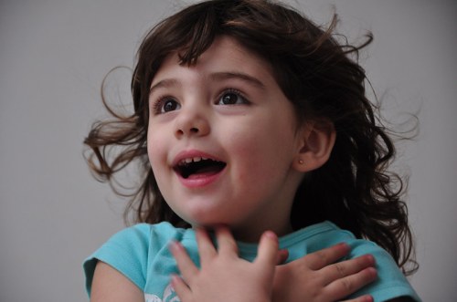 A joyful young girl with curly hair smiles widely, her hands clasped near her chest, against a neutral background.