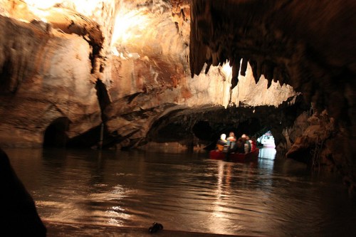A boat with people navigates through a dark, rocky cave with water reflecting the light from above.